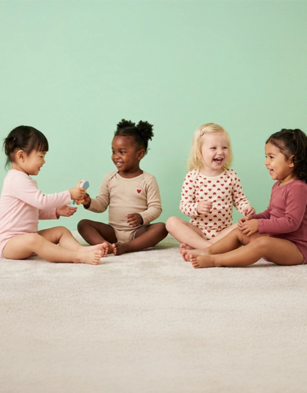 Een groep van vier peuters zit op een lichte vloer en speelt samen met speelgoed voor een effen pastelgroene achtergrond.