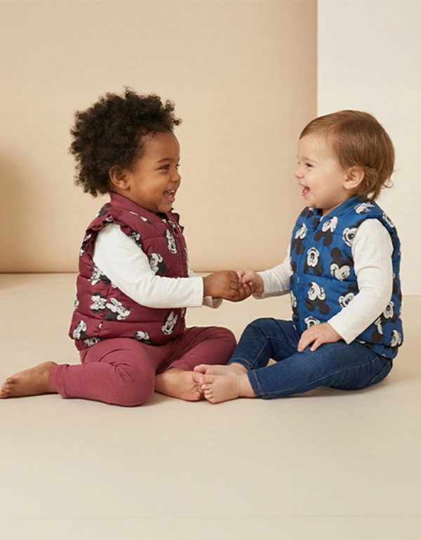 Twee peuters zitten voor een lichte achtergrond, dragen kleurrijke vesten, houden elkaars hand vast en lachen elkaar toe.
