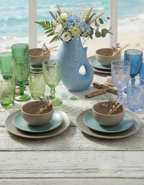 Maritiem gedekte tafel met kleurrijke glazen, servies en decoratieve vaas met bloemen tegen zeezicht – stijlvolle tafeldecoratie voor zomerse momenten