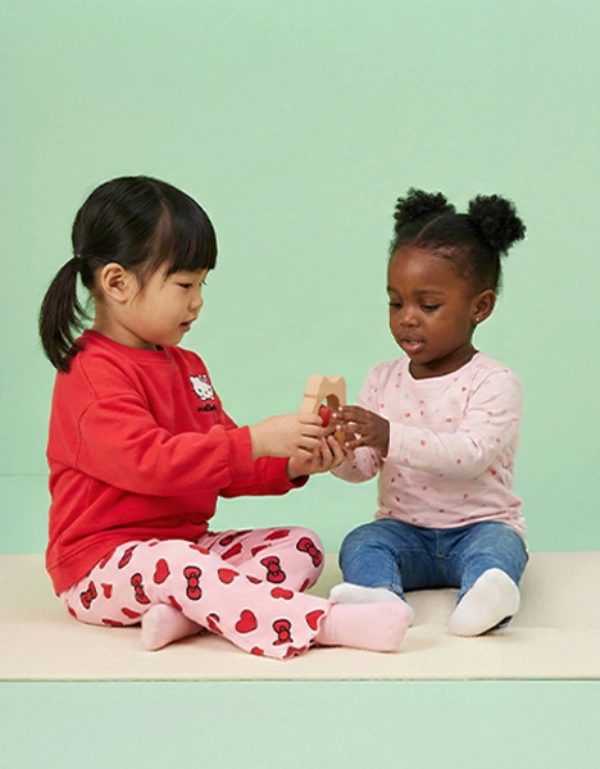 Twee kleine kinderen zitten op een zachte ondergrond en spelen vrolijk samen, terwijl ze kleurrijk speelgoed vasthouden tegen een pastelgroene achtergrond.