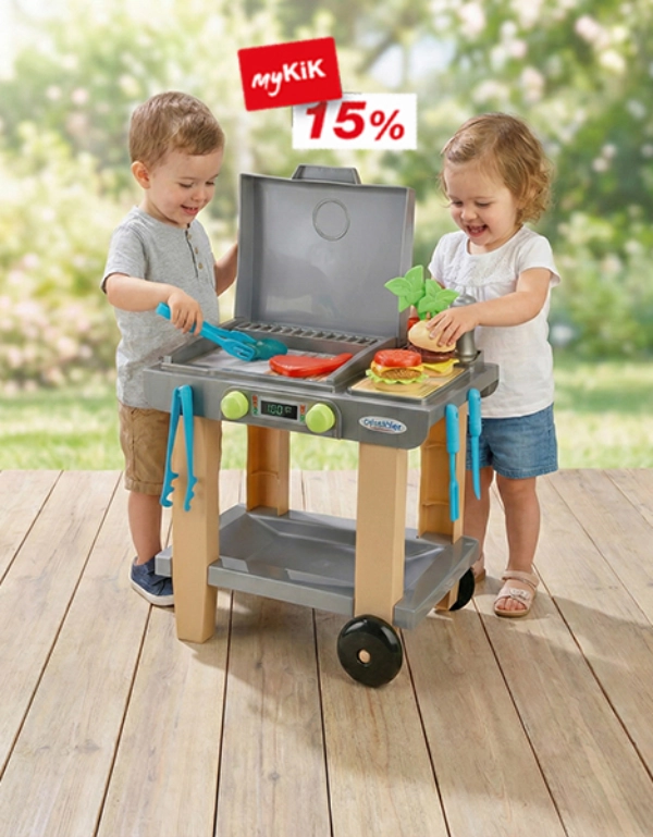 Twee kinderen spelen met een speelgoedgrill, met kleurrijke keukengerei en nepvoedsel, op een houten terras omgeven door groen.