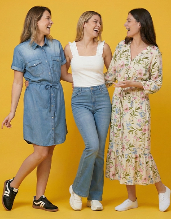 Drie vrouwen staan samen tegen een felgele achtergrond, elk gekleed in een stijlvolle outfit: een spijkerjurk, een casual top met jeans en een maxi-jurk met bloemenprint.