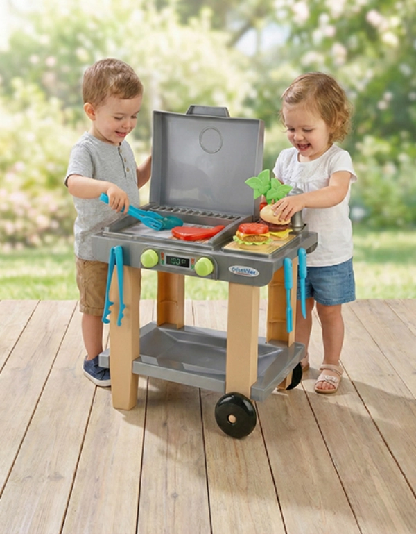 Twee kinderen spelen met een speelgoedgrill, met kleurrijke keukengerei en nepvoedsel, op een houten terras omgeven door groen.