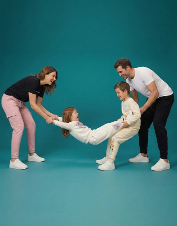 Een gezin speelt speels met elkaar en houdt een kind tussen twee volwassenen; allemaal casual gekleed tegen een turquoise achtergrond.