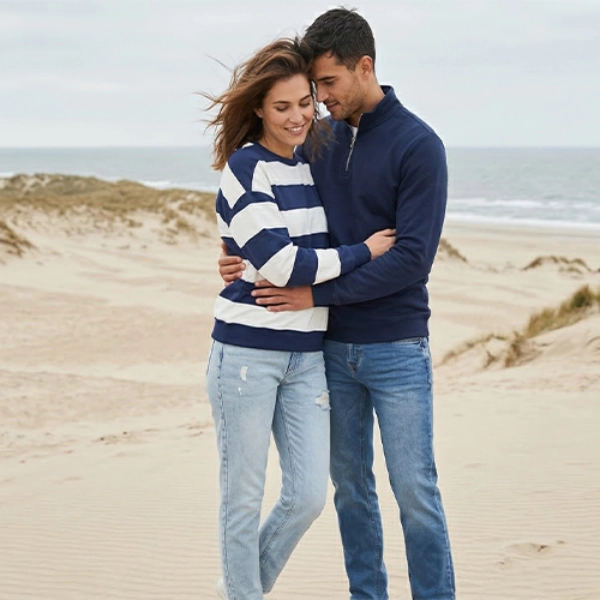 Een stel in casual kleding omarmt elkaar op een zandstrand met duinen en oceaangolven onder een bewolkte lucht.