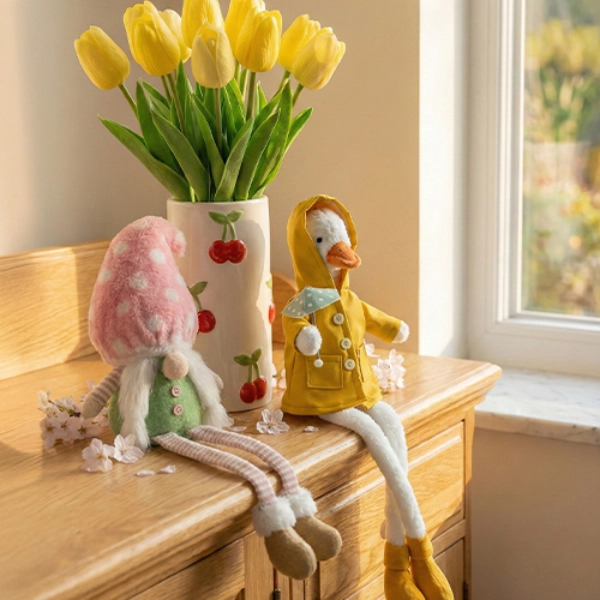 Twee knuffelfiguren zitten op een houten oppervlak naast een vaas met felgele tulpen. Het tafereel is vrolijk en kleurrijk.