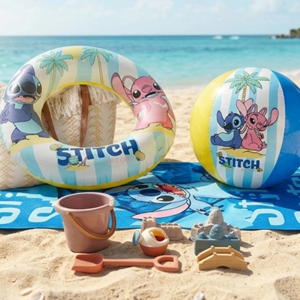 Strandhanddoek, zwemring, bal en strandspeelgoed met Lilo & Stitch‑thema liggen op het zandstrand naast de turquoise zee.