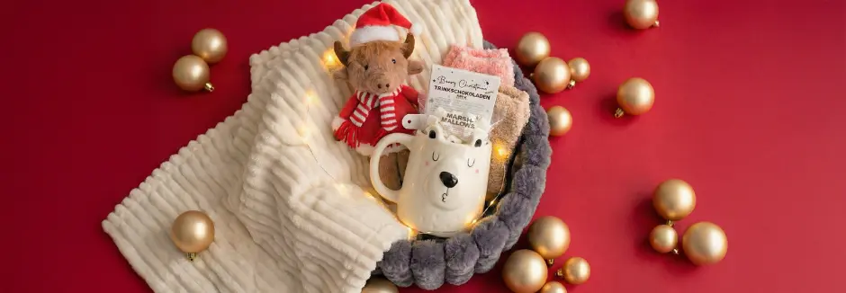 Een gezellig kersttafereel met een pluchen rendier, een berenmok met marshmallows, warme sokken en gouden ornamenten op een rode achtergrond.