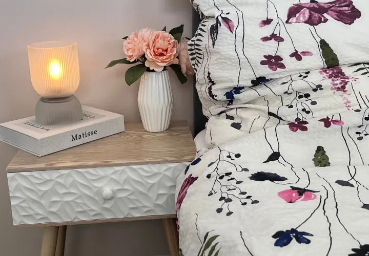 Een knus tafereel naast het bed met een brandende lamp, een bloemenvaas met roze rozen en een dekbed met patroon tegen een zachte, neutrale muur.