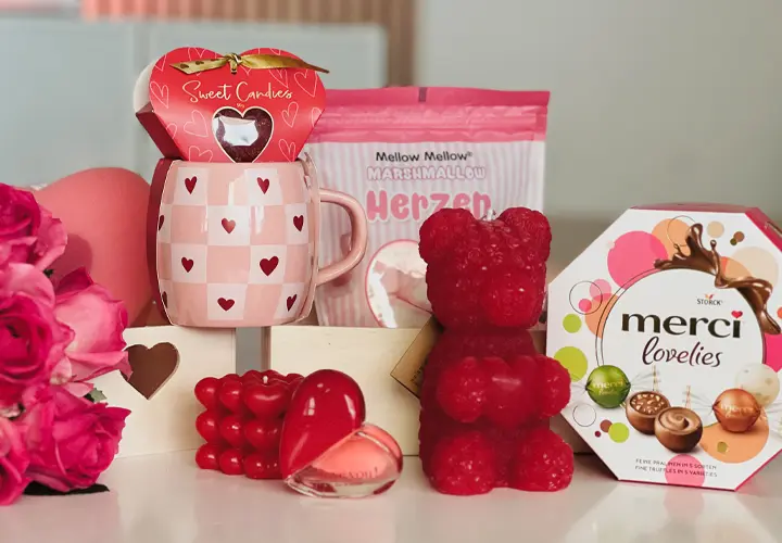 Een feestelijke opstelling van Valentijnscadeaus: een mok met hartjespatroon, snoepjes, een rode gummybeer en een doos chocolaatjes, omlijst door rozen.