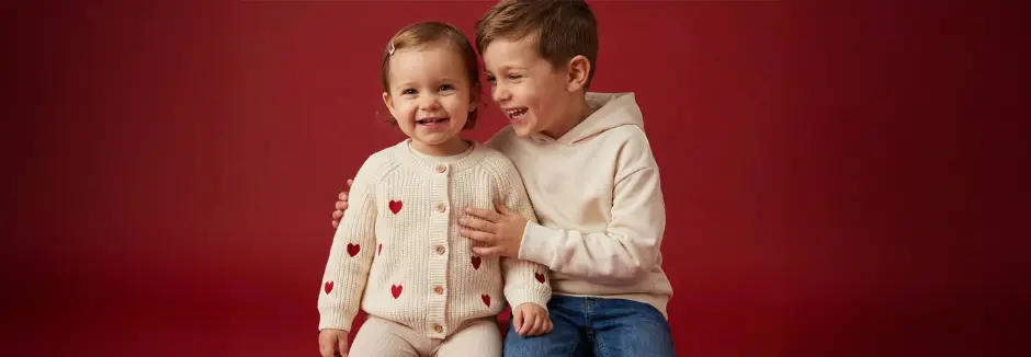 Twee kinderen zitten naast elkaar, dragen comfortabele kleding en glimlachen voor een dieprode achtergrond. Het meisje draagt een crèmekleurig vest met rode hartjes.