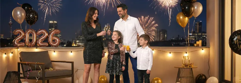 Een gezin viert feest met drankjes op een balkon, terwijl vuurwerk de nachtelijke hemel achter hen verlicht.