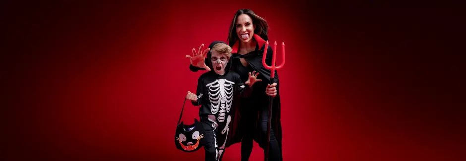 Een kind in een skeletkostuum houdt een zwarte tas met kattenmotief vast, terwijl een volwassene in een duivelspak met een drietand poseert tegen een rode achtergrond.