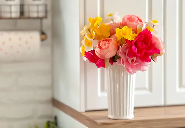 Kleurrijke kunstbloemen in een witte vaas, uitgestald op een houten plank tegen een lichte keukenachtergrond.