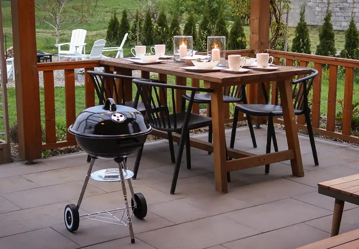 Een gezellig eetgedeelte buiten met een houten tafel met kaarsen, stoelen en een kleine zwarte barbecue ernaast, midden in een groene tuin.