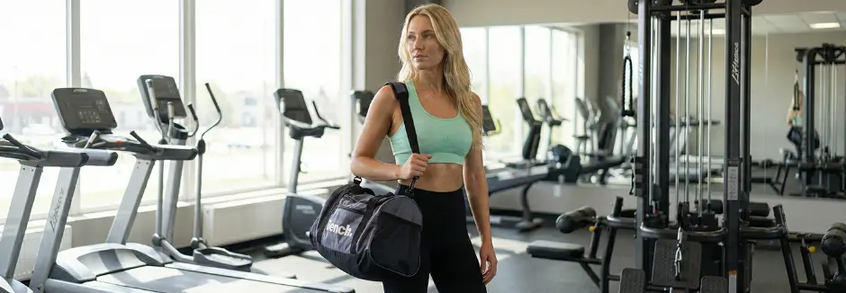 Eine selbstbewusste Person steht in einem Fitnessstudio und hält eine Sporttasche, während Trainingsgeräte und große Fenster im Hintergrund zu sehen sind.