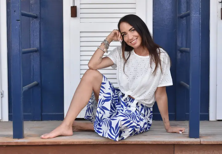 Een vrouw zit gracieus op een veranda, draagt een wit topje en een blauwe gebloemde rok, met haar hand rustend op haar kin, diep in gedachten.