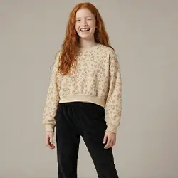 Meisje draagt een beige gebreide trui en zwarte broek tegen een lichte achtergrond.