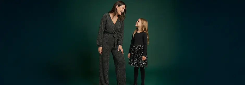 Eine Frau in einem glitzernden schwarzen Overall steht neben einem Mädchen in einem schwarzen Oberteil mit einem Paillettenrock, beide lächeln vor einem dunkelgrünen Hintergrund.