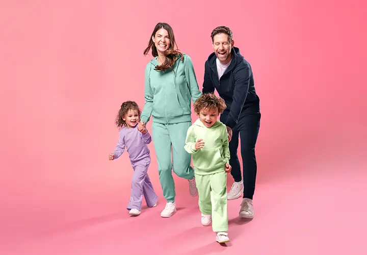 Een gezin van vier in kleurrijke bijpassende outfits loopt vrolijk hand in hand tegen een zachtroze achtergrond.