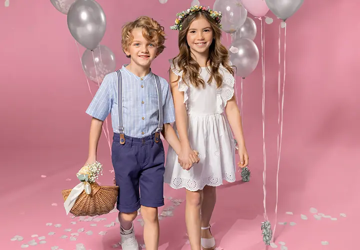 Twee kinderen houden elkaars hand vast op een roze achtergrond. De een draagt een gestreept shirt en een korte broek, de ander een witte jurk en een bloemenkroon.