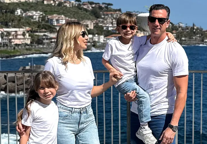 Een gezin van vier poseert samen aan zee, gekleed in bijpassende witte T-shirts en jeans, met een schilderachtig decor van heuvels en water op de achtergrond.