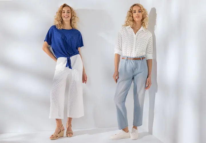 Twee vrouwen modelleren stijlvolle outfits: de ene in een blauwe top en witte broek, de andere in een witte blouse en lichtblauwe broek tegen een lichte achtergrond.