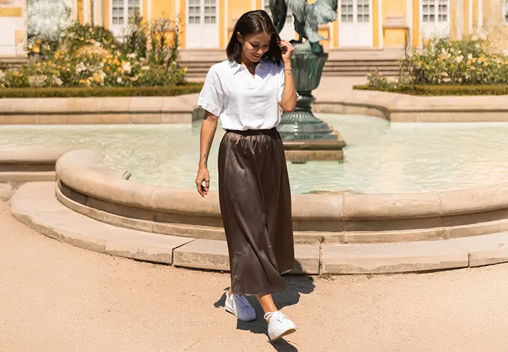 Een vrouw in een wit shirt en een bruine rok loopt langs een fontein omringd door groen, met gele gebouwen op de achtergrond.