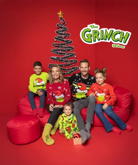 Een feestelijk familieportret met vijf leden in kleurrijke truien met Grinch-thema, zittend op rode zitzakken, voor een kerstboom.
