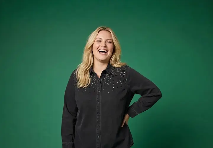 Een vrouw met golvend blond haar staat zelfverzekerd in een zwart shirt met subtiele versieringen tegen een groene achtergrond.