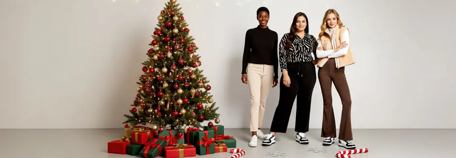 Drie vrouwen poseren samen bij een prachtig versierde kerstboom met cadeautjes, waardoor een feestelijke kerstsfeer ontstaat.