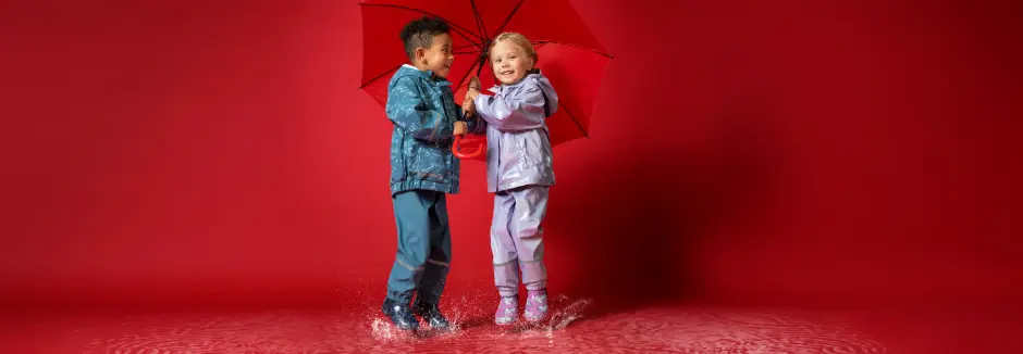Twee kinderen in felgekleurde regenjassen spelen vrolijk in plassen terwijl ze een felrode paraplu delen.