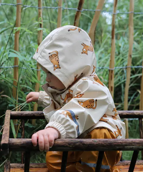 Ein Kind spielt in der regnerischen Natur und hat eine beige Regenjacke mit Katzenmotiven und die dazugehörige braune Matchhose an 