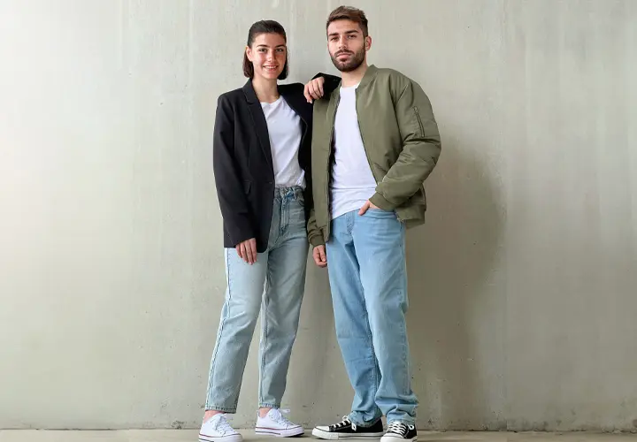 Een man en een vrouw staan zij aan zij voor een eenvoudige muur, allebei gekleed in vrijetijdskleding met jeans en sneakers.