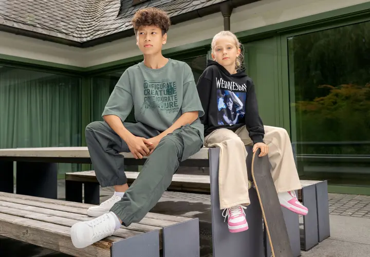 Een jongen in een oversized groen T-shirt en groene broek zit op een houten bank naast een meisje in een zwarte hoodie en beige broek, met een skateboard in haar hand.