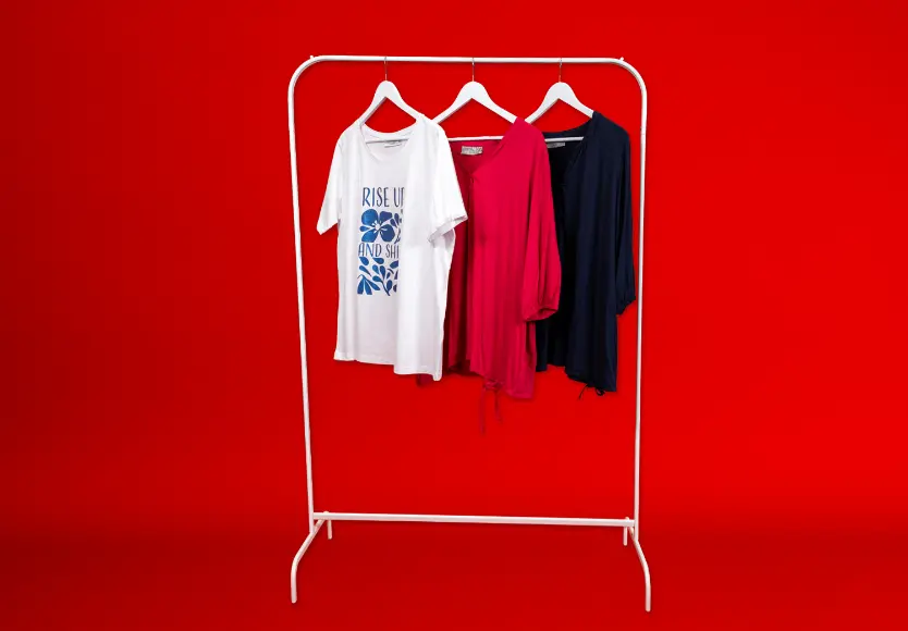 Drie stijlvolle tops hangen aan een wit kledingrek tegen een felrode achtergrond: een wit T-shirt met opdruk, een roze blouse en een marineblauwe blouse.