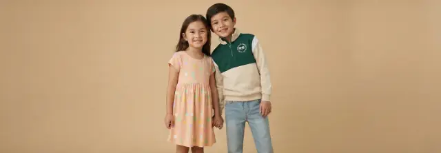Twee kinderen staan samen tegen een effen beige achtergrond. Het meisje draagt een kleurrijke jurk, terwijl de jongen een groen en crèmekleurig sweatshirt draagt.