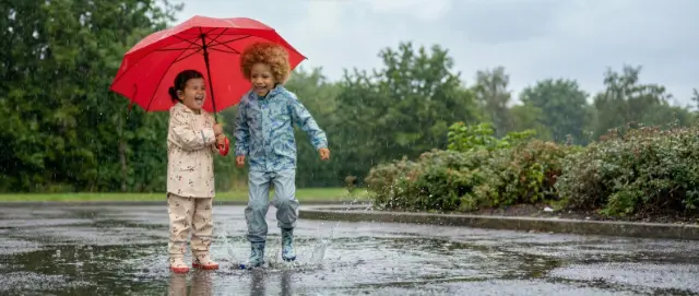 Droog door de regenachtige dag Twee kinderen spelen in de regen, spetteren in plassen en delen een knalrode paraplu, omringd door veel groen.
