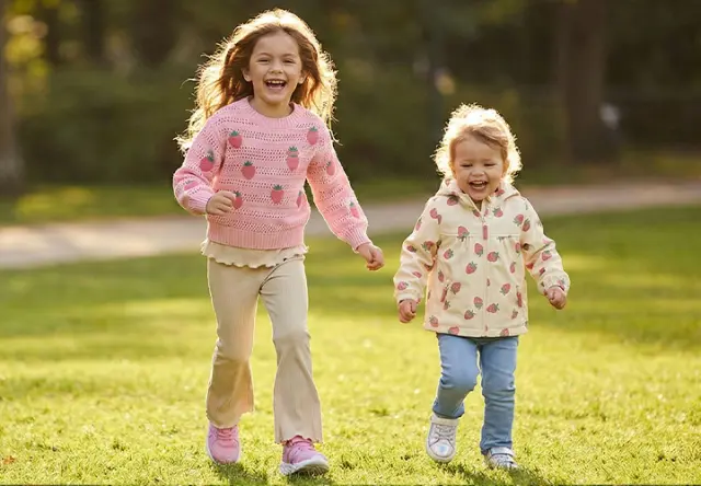 Twee kinderen lopen samen door een zonnig park. Ze dragen kleurrijke kleding met fruitpatronen en genieten van een vrolijk moment buiten.