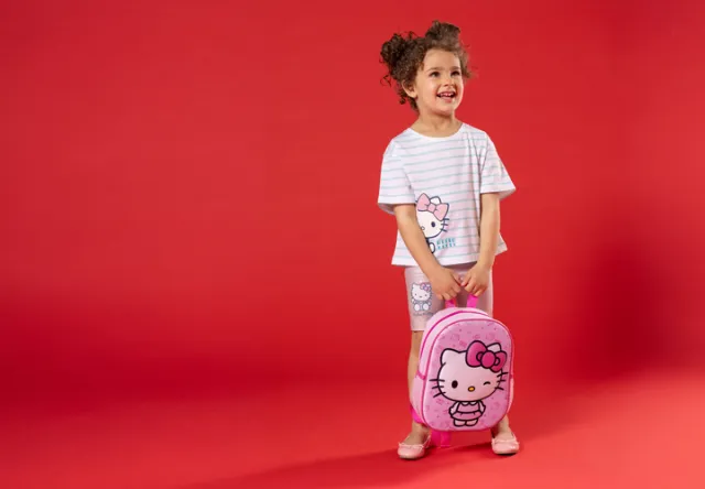 Een kind staat voor een rode achtergrond, gekleed in een gestreept Hello Kitty-shirt en korte broek, met een roze Hello Kitty-rugzak in de hand.