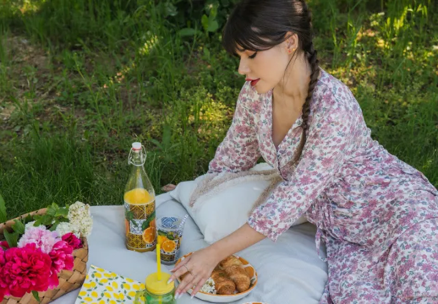Een picknickscène met een vrouw in een bloemenjurk, omringd door kleurrijke bloemen, sinaasappeldrank, croissants en een kleurrijke deken op het gras.