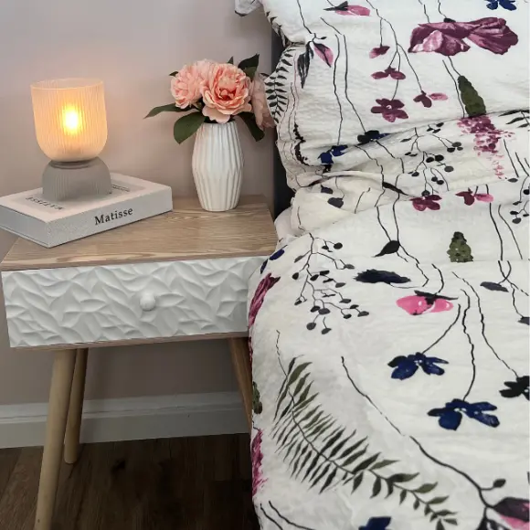 Een knus tafereel naast het bed met een brandende lamp, een bloemenvaas met roze rozen en een dekbed met patroon tegen een zachte, neutrale muur.