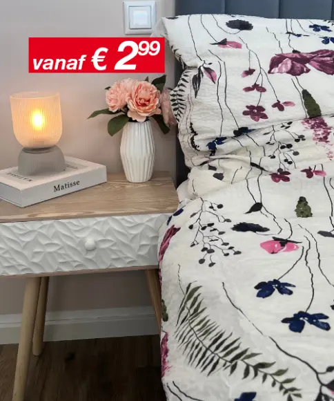 Een knus tafereel naast het bed met een brandende lamp, een bloemenvaas met roze rozen en een dekbed met patroon tegen een zachte, neutrale muur.