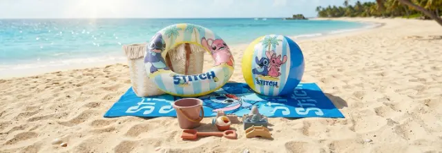 Strandhanddoek, zwemring, bal en strandspeelgoed met Lilo &amp; Stitch‑thema liggen op het zandstrand naast de turquoise zee.