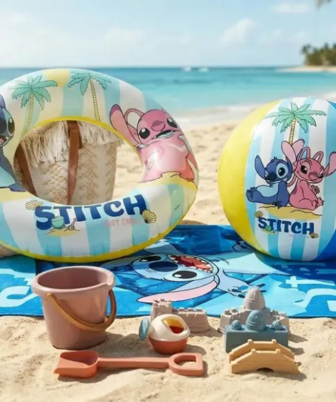 Strandhanddoek, zwemring, bal en strandspeelgoed met Lilo & Stitch‑thema liggen op het zandstrand naast de turquoise zee.