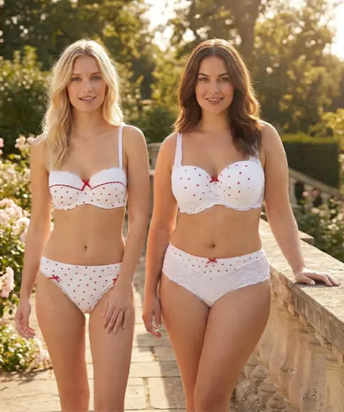 Twee vrouwen staan op een stenen pad, omringd door bloeiende roze rozen, en dragen bijpassend wit ondergoed met hartjes in een zonovergoten tuin.