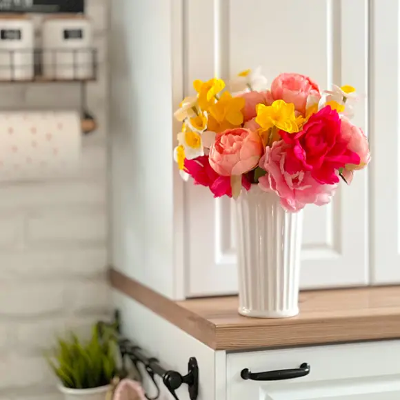 Kleurrijke kunstbloemen in een witte vaas, uitgestald op een houten plank tegen een lichte keukenachtergrond.