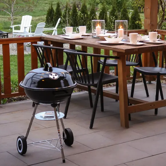 Een gezellig eetgedeelte buiten met een houten tafel met kaarsen, stoelen en een kleine zwarte barbecue ernaast, midden in een groene tuin.