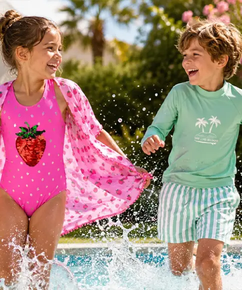 Twee kinderen rennen bij het zwembad, één draagt een roze badpak met aardbeienprint en een roze strandjurkje, de ander een groen shirt met lange mouwen met palmprint en groen‑wit gestreepte zwemshort.