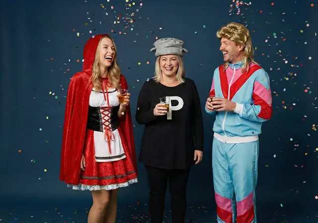 Drie vrienden in kostuums vieren feest met drankjes, omringd door kleurrijke confetti tegen een blauwe achtergrond.